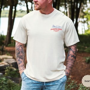Puede incluir: Una camiseta de manga corta color marfil con un peque&ntilde;o gr&aacute;fico en la parte superior izquierda del pecho. El gr&aacute;fico muestra el contorno azul de unas monta&ntilde;as sobre el texto rojo "JOHNSON FAMILY Vacation 2026". Tiene cuello redondo y corte est&aacute;ndar.