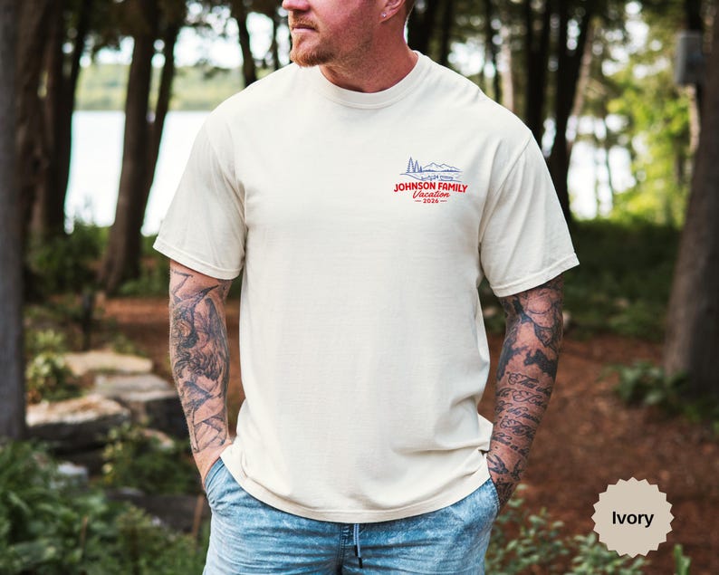 Puede incluir: Una camiseta de manga corta color marfil con un peque&ntilde;o gr&aacute;fico en la parte superior izquierda del pecho. El gr&aacute;fico muestra el contorno azul de unas monta&ntilde;as sobre el texto rojo "JOHNSON FAMILY Vacation 2026". Tiene cuello redondo y corte est&aacute;ndar.