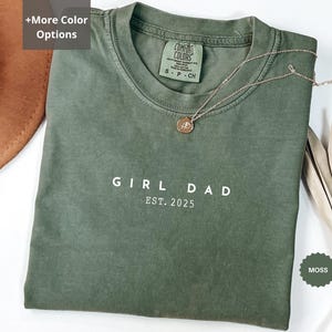 Pode incluir: Uma camiseta verde musgo com as palavras "GIRL DAD EST. 2025" impressas em branco. A camiseta tem gola redonda e mangas curtas. Um colar de ouro com um pingente repousa sobre a camiseta. A camiseta est&aacute; dobrada sobre uma superf&iacute;cie branca.