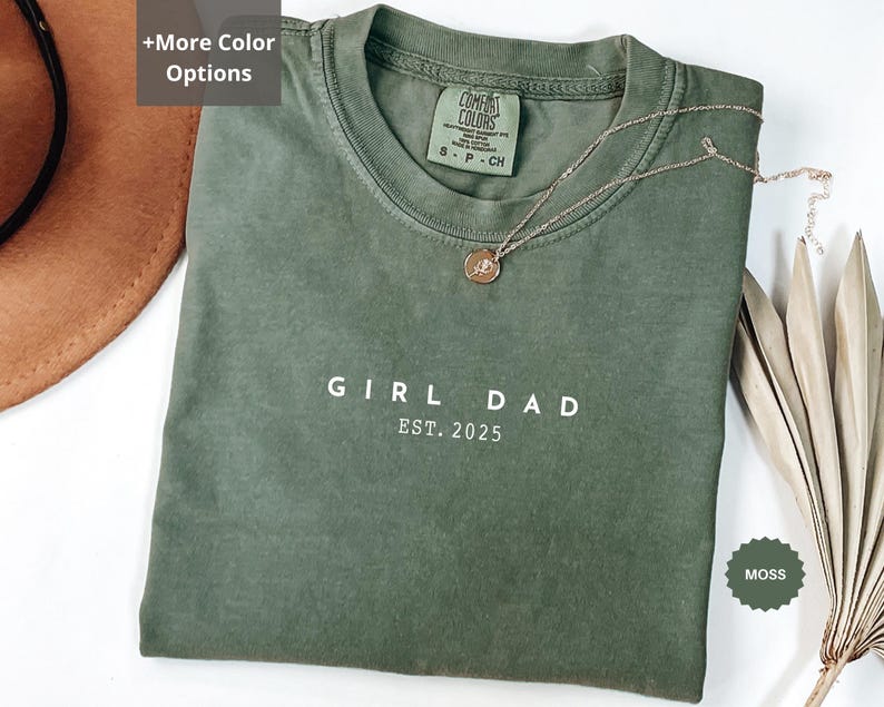 Pode incluir: Uma camiseta verde musgo com as palavras "GIRL DAD EST. 2025" impressas em branco. A camiseta tem gola redonda e mangas curtas. Um colar de ouro com um pingente repousa sobre a camiseta. A camiseta est&aacute; dobrada sobre uma superf&iacute;cie branca.