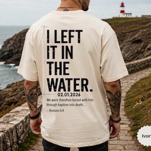 Puede incluir: Camiseta color marfil con el texto negro "I LEFT IT IN THE WATER", la fecha 02.01.2026 y un versículo bíblico. La camiseta es de corte holgado, con un gráfico de cruz encima del texto.