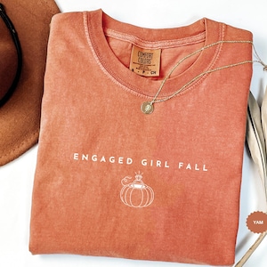 May include: A terracotta-colored short-sleeve t-shirt with a crew neckline. The shirt features the white text "ENGAGED GIRL FALL" above a white line-art graphic of a pumpkin with a diamond ring on top. The interior label reads "COMFORT COLORS".