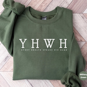 Puede incluir: Una sudadera verde militar con texto blanco "YHWH" sobre las palabras "EVERY BREATH SPEAKS HIS NAME". La sudadera tiene cuello redondo y mangas largas con puños acanalados. El fondo es un patrón de madera marrón claro.
