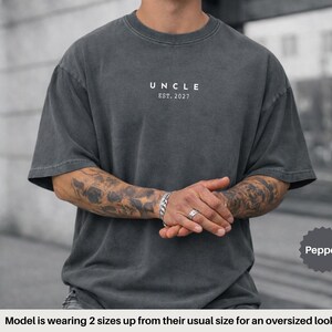May include: A charcoal grey oversized t-shirt featuring the white text UNCLE EST. 2027 printed on the chest. The garment has a crew neckline and short sleeves. A small circular badge in the bottom right corner contains the word Pepper.