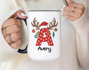 May include: White ceramic mug with a black rim and handle. The mug features a red letter "A" with white polka dots, adorned with reindeer antlers, a Santa hat, and Christmas ornaments. The name "Avery" is written below.