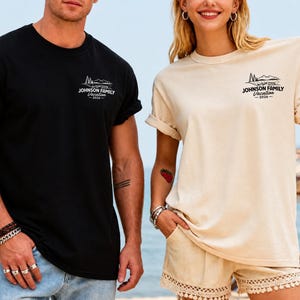 以下が含まれることがあります： 「Johnson Family Vacation 2026」の文字と山のグラフィックが入った黒とベージュのTシャツ。Tシャツは袖がまくられています。背景にはビーチの風景が写っています。