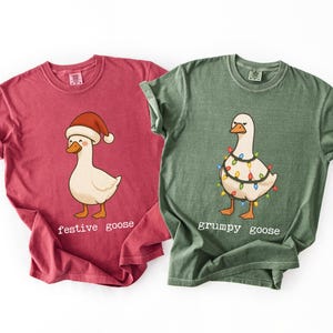 Puede incluir: Una camiseta roja con una oca de dibujos animados con un gorro de Papá Noel y el texto 'festive goose' y una camiseta verde con una oca envuelta en luces navideñas de colores con el texto 'grumpy goose' impreso debajo de los gráficos.