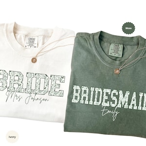 May include: Two t-shirts, one ivory and one moss green, with the words "BRIDE" and "BRIDESMAID" in a floral design. Each shirt has a gold necklace with a pendant. The ivory shirt says "Mrs. Johnson" and the green shirt says "Emily".