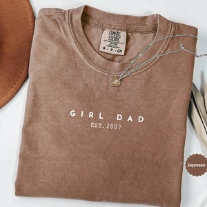 Pode incluir: Camiseta cor de caf&eacute; com o texto branco "GIRL DAD EST. 2007". Um colar de prata com um pingente repousa sobre a camiseta. Um chap&eacute;u marrom e folhas secas tamb&eacute;m est&atilde;o na imagem.