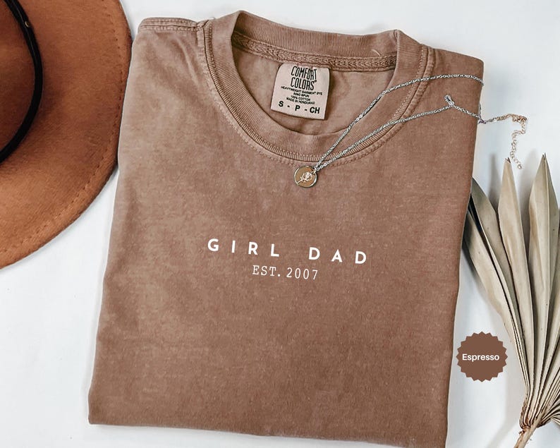 Pode incluir: Camiseta cor de caf&eacute; com o texto branco "GIRL DAD EST. 2007". Um colar de prata com um pingente repousa sobre a camiseta. Um chap&eacute;u marrom e folhas secas tamb&eacute;m est&atilde;o na imagem.