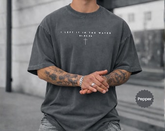 May include: A charcoal gray short-sleeve t-shirt with a crew neckline. The chest features white text that reads I LEFT IT IN THE WATER, followed by the date 01.03.26 and a small cross symbol centered below the text. The fabric has a washed finish.