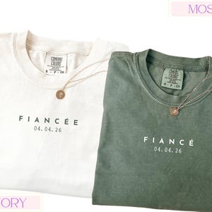Puede incluir: Dos camisetas Comfort Colors, una marfil y otra verde musgo, con la palabra "FIANC&Eacute;E" y la fecha "04.04.26" impresas en la parte delantera. Cada camiseta tiene un collar con colgante dorado. Las camisetas son talla peque&ntilde;a.