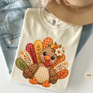 May include: An ivory t-shirt featuring a graphic of a turkey with patterned feathers in shades of orange, brown, green, and yellow. The turkey has a floral crown on its head and a gingham chest. The shirt has a Comfort Colors label at the neck.