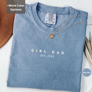 Pode incluir: Uma camiseta azul jeans com o texto "GIRL DAD EST. 2026" impresso em branco. Um colar de ouro com um pingente repousa sobre a camisa. A camisa tem gola redonda e uma etiqueta pequena.