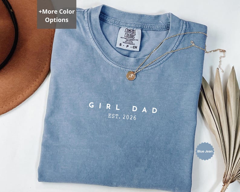 Pode incluir: Uma camiseta azul jeans com o texto "GIRL DAD EST. 2026" impresso em branco. Um colar de ouro com um pingente repousa sobre a camisa. A camisa tem gola redonda e uma etiqueta pequena.