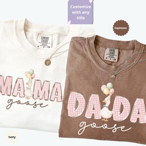 May include: Two short-sleeve t-shirts in ivory and espresso colors. The ivory shirt features the text MAMA goose with a goose wearing a party hat and balloons. The espresso shirt features the text DADA goose with a goose wearing a party hat and balloons. Both shirts have pink gingham lettering.