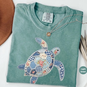 May include: Light green t-shirt with a patchwork turtle design. The turtle is made of various blue floral and gingham fabrics. A gold necklace with a small pendant rests on the shirt. The shirt has a Comfort Colors tag.