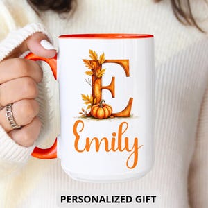 May include: White ceramic mug with an orange interior and handle. The mug features a fall-themed design with the letter "E" decorated with leaves and a pumpkin, and the name "Emily" in orange script. The words "PERSONALIZED GIFT" are at the bottom.