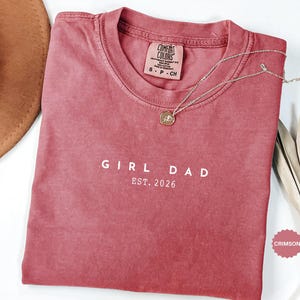 Pode incluir: Camiseta carmesim com o texto branco "GIRL DAD EST. 2026". Um colar dourado com um pingente repousa sobre a camisa. A camisa tem gola redonda e uma etiqueta Comfort Colors. Um chap&eacute;u marrom &eacute; parcialmente vis&iacute;vel.