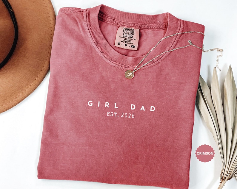 Pode incluir: Camiseta carmesim com o texto branco "GIRL DAD EST. 2026". Um colar dourado com um pingente repousa sobre a camisa. A camisa tem gola redonda e uma etiqueta Comfort Colors. Um chap&eacute;u marrom &eacute; parcialmente vis&iacute;vel.