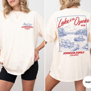 Puede incluir: Una camiseta color marfil con un peque&ntilde;o gr&aacute;fico en el pecho y un gr&aacute;fico grande en la espalda. El dise&ntilde;o trasero muestra una escena de lago con dos sillas en un muelle, un barco y el texto Lake of the Ozarks 2026 y JOHNSON FAMILY Vacation.