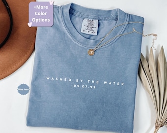 May include: A blue jean Comfort Colors t-shirt with the text "WASHED BY THE WATER 09.07.25" in white. A gold necklace with a pendant is draped across the shirt. The shirt is folded and placed on a white surface.