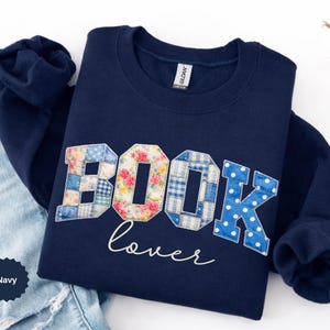 May include: Navy blue sweatshirt with the word "BOOK" in patchwork letters and the word "lover" in cursive. The patchwork includes floral, plaid, and polka dot patterns. The sweatshirt is folded on top of denim shorts.