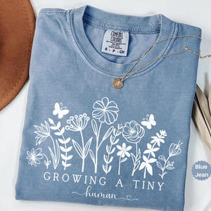 May include: A blue jean-colored t-shirt with a white floral design and the text "GROWING A TINY human". A gold necklace with a pendant is draped across the shirt. The shirt is folded and has a Comfort Colors tag.