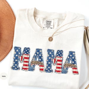 May include: Ivory-colored Comfort Colors sweatshirt with the word "MAMA" appliqued across the front. The letters feature a patchwork design with American flag and leopard print patterns. A gold necklace and a brown hat are also in the image.