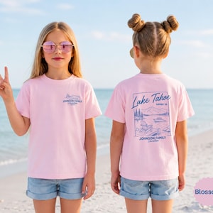 以下が含まれることがあります： 浜辺に立っている、明るいピンクのTシャツとデニムショーツを着た2人の若い人たち。シャツの前面には小さな青いグラフィックとテキストがあります。シャツの背面には、テキスト付きのレイクタホのデザインが施されています。シャツの色はBlossomと呼ばれています。