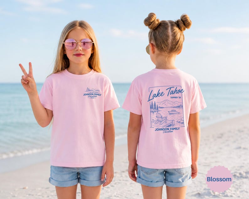 以下が含まれることがあります： 浜辺に立っている、明るいピンクのTシャツとデニムショーツを着た2人の若い人たち。シャツの前面には小さな青いグラフィックとテキストがあります。シャツの背面には、テキスト付きのレイクタホのデザインが施されています。シャツの色はBlossomと呼ばれています。