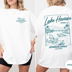 以下が含まれることがあります： 白のTシャツ、ターコイズブルーのグラフィック。前面には「JOHNSON FAMILY Vacation - 2026」の文字。背面には「Lake Havasu SUMMER '26」と湖の風景、ボート、そして「JOHNSON FAMILY Vacation」の文字が描かれています。