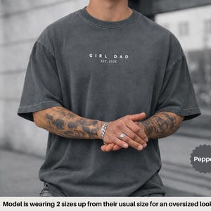 Pode incluir: T-shirt oversized cor pimenta com o texto "GIRL DAD EST. 2026" em branco. A camisa tem gola redonda e mangas curtas. A modelo est&aacute; vestindo a camisa dois tamanhos acima para um look oversized.