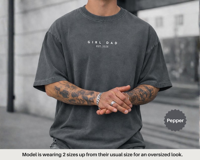 Pode incluir: T-shirt oversized cor pimenta com o texto "GIRL DAD EST. 2026" em branco. A camisa tem gola redonda e mangas curtas. A modelo est&aacute; vestindo a camisa dois tamanhos acima para um look oversized.
