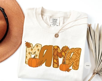 May include: Ivory-colored Comfort Colors t-shirt with the word "MAMA" in a fall-themed design. The letters are filled with patterns of acorns, leaves, and flowers. Two orange pumpkins and leaves are below the word. A gold necklace is draped on the shirt.