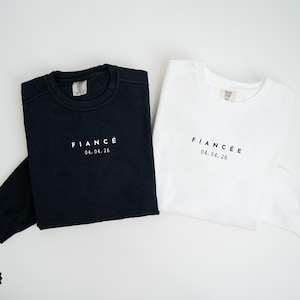 Puede incluir: Se muestran dos sudaderas de manga larga, una negra y otra blanca. La sudadera negra tiene la palabra "FIANC&Eacute;" y la fecha "04.04.26" impresas en blanco. La sudadera blanca tiene el mismo texto en negro.