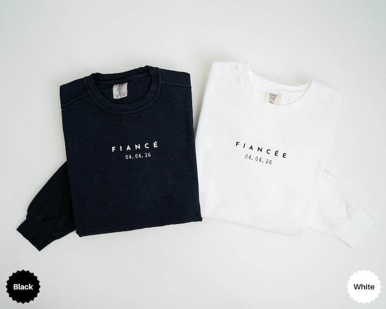 Puede incluir: Se muestran dos sudaderas de manga larga, una negra y otra blanca. La sudadera negra tiene la palabra "FIANC&Eacute;" y la fecha "04.04.26" impresas en blanco. La sudadera blanca tiene el mismo texto en negro.