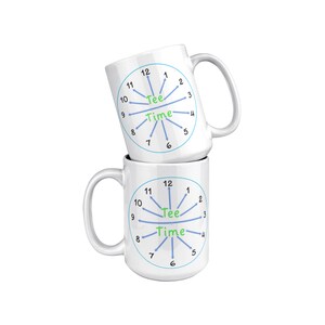 Puede incluir: Dos tazas de cerámica blanca apiladas una encima de la otra. Ambas tazas presentan un círculo azul con un diseño de esfera de reloj. La esfera del reloj tiene líneas azules que irradian desde el centro hasta los números del 1 al 12. Las palabras "Tee Time" están escritas en verde en el centro de la esfera del reloj.