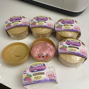 Puede incluir: Cinco cestas de vapor redondas de plástico color canela con juguetes de masa blandita. Cada cesta tiene una etiqueta que dice The Original Mystery Squishy Dumpling The Viral Fidget Toy. Una cesta está abierta y muestra una masa rosa sonriente.