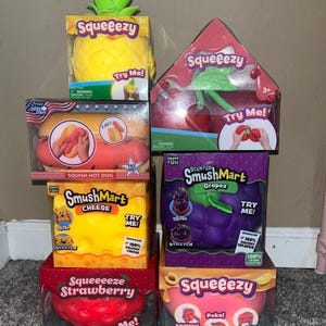Könnte beinhalten: Eine Sammlung von sechs Squeezy- und SmushMart-Quetschspielzeugen in Lebensmittelform in ihrer Originalverpackung. Die Artikel umfassen eine Ananas, einen Hotdog, einen Käseblock, eine Weintraube, eine Erdbeere und einen Pfirsich in bunten Farben.