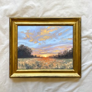 以下が含まれることがあります： 夕暮れの草原を描いた額装された絵画。空はふわふわの雲のある鮮やかな青色で、太陽は木々の列の後ろに沈んでいます。草原は金色で、夕暮れの光が長い影を落としています。