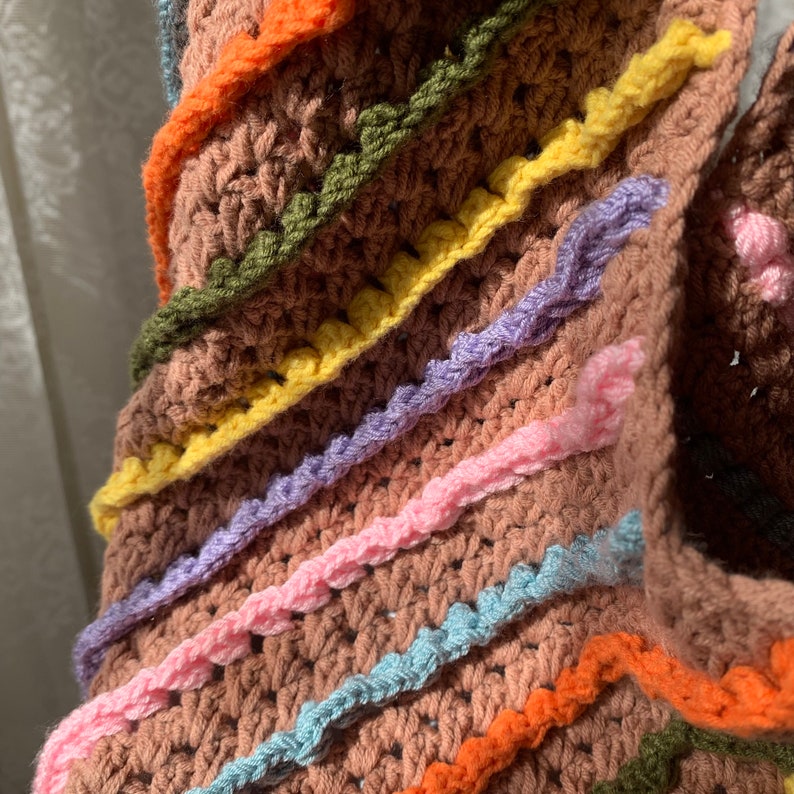 May include: Close-up of a brown crocheted blanket with colourful stripes of pink, blue, yellow, green, and orange. The blanket has a textured, bumpy surface.