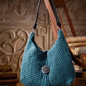 May include: A teal blue crocheted handbag with a silver chain strap and a decorative button with a tassel. The bag is hanging on a wooden chair.