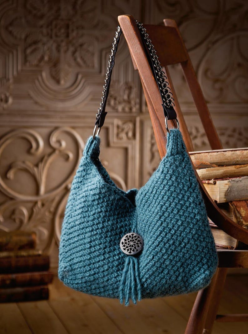 May include: A teal blue crocheted handbag with a silver chain strap and a decorative button with a tassel. The bag is hanging on a wooden chair.