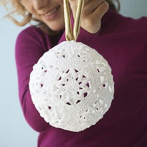 May include: A white crocheted bauble with a delicate snowflake pattern. The bauble is being held by a person's hand.