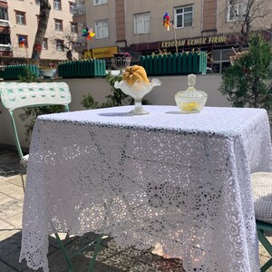 May include: A white, crocheted lace tablecloth covers a small outdoor table. Two glass serving dishes and a vintage metal chair are also visible. The table is set outside on a sunny day.