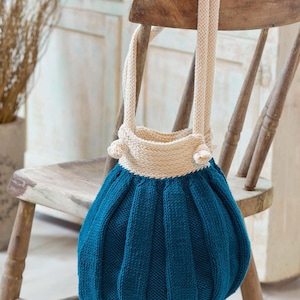 May include: A teal blue knitted tote bag with a cream colored top and straps. The bag is hanging on a wooden chair.