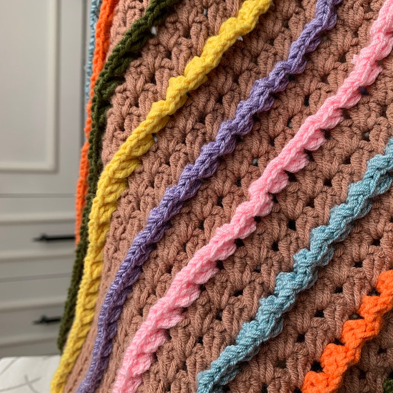 May include: A close-up of a crocheted blanket with a rainbow pattern. The blanket is made with different coloured yarns, including yellow, purple, pink, blue, and orange. The blanket is crocheted in a textured stitch, creating a raised pattern.