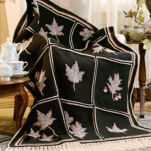 Puede incluir: Una manta de crochet negra con borde de flecos presenta un patrón repetido de hojas de arce de colores.