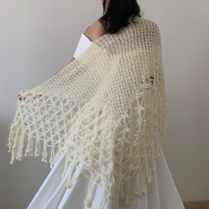 May include: A white crocheted shawl with a delicate floral pattern and fringe. The shawl is draped over a white dress.
